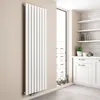 A vertical designer radiator is shown in a modern kitchen setting, next to a large window. Produktbild-Vorschau