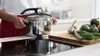 A person is holding a pressure cooker on a stovetop, with vegetables on a cutting board nearby. Anteprima dell’immagine prodotto