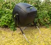 A black compost tumbler sits on a stand in a garden setting, surrounded by green bushes and fallen yellow leaves. Náhled obrázku produktu