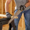 A person wearing work gloves is using a handsaw to cut a piece of wood. The wood is supported by a black sawhorse. The background is a wooden wall. Náhled obrázku produktu