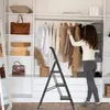A woman is organizing clothes in a closet, using a black stepladder. Produktbild-Vorschau