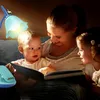A mother is reading a book to her two young children in bed at night, illuminated by a bedside lamp. Produktbild-Vorschau
