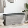 A storage bench with a gray upholstered top and a light wood-grain base, placed in an entryway with a coat hanging on the wall and a plant in the background. Náhled obrázku produktu