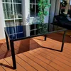 A black glass-top dining table with a metal frame sits on a wooden deck. Produktbild-Vorschau