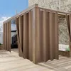 Outdoor curtains hanging from a wooden pergola structure on a patio with a stone wall in the background. Produktbild-Vorschau