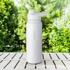 A white water bottle is placed on a wooden table outdoors with green foliage in the background. Produktbild-Vorschau