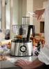 A person's hands are adding liquid to a food processor containing ingredients, with other ingredients and baking supplies scattered on the counter. Anteprima dell’immagine prodotto