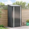 A dark gray metal garden shed is shown in an outdoor setting, placed on a paved patio next to a wooden fence. A potted plant and a green watering can are visible to the right of the shed. Produktbild-Vorschau