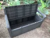 An open dark gray plastic storage bench is placed outdoors on gravel, surrounded by lush green plants. Produktbild-Vorschau