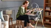 A woman is sitting on a stool next to a baby bouncer/high chair with a baby in it. The scene is indoors, likely a living room or nursery. Náhled obrázku produktu
