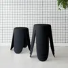 Two black stools are placed on a white floor in front of a black and white grid patterned wall. A green plant hangs from the top right corner. Produktbild-Vorschau