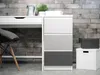 A close-up of a white desk with a drawer, a three-drawer cabinet with varying shades of gray drawers, a chair with a gray cushion, a watering can, a potted plant, and storage boxes against a brick wall. Produktbild-Vorschau
