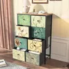 A multi-drawer cabinet with decorative fabric fronts, placed in a room setting with curtains and a lamp. Náhled obrázku produktu