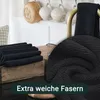 A stack of black microfiber cloths is shown on a wooden surface, with a close-up view of the fabric texture in the foreground. In the background, mugs hang from hooks above a white shelf. Produktbild-Vorschau