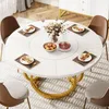 A round white dining table with a gold base, set with placemats, plates, cutlery, a fruit bowl, and a carafe of red wine, surrounded by chairs in a dining room setting. Podgląd zdjęcia produktu