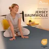 A woman sitting on a bed, smiling, with a yellow pillow and a bedside table with plants and a lamp in the background. Produktbild-Vorschau