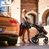 A woman is pushing a stroller next to a luxury car in front of a modern building. Produktbild-Vorschau