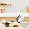 A white and black hand mixer sits on a wooden countertop, with desserts and whipped cream in the foreground. Produktbild-Vorschau