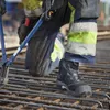A construction worker is kneeling on rebar, using bolt cutters. The focus is on the worker's boot and lower leg. Produktbild-Vorschau