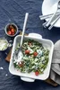 A close-up overhead shot of a pasta dish in a white baking dish, with a serving utensil, small bowls of cherry tomatoes and grated cheese, and cutlery on a dark blue tablecloth. Podgląd zdjęcia produktu