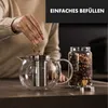 A person is adding tea leaves to a glass teapot, with a jar of tea leaves next to it. Produktbild-Vorschau