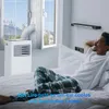 A man relaxing in bed in a bedroom, with a portable air conditioner unit set up by the window. Produktbild-Vorschau