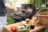 A person is grilling food on a BBQ grill in a backyard setting. A table in the foreground is laden with fresh vegetables, drinks, and a basket. Podgląd zdjęcia produktu