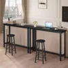 A long, narrow black bar table is placed against a wall with a sofa behind it. Two black bar stools are positioned in front of the table. A laptop, a cup, and a small vase of flowers are on the table. Náhľad obrázku produktu