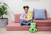 A young girl sits on a pink modular sofa, holding two plush teddy bears. A third plush teddy bear sits on the floor in front of her. A potted plant is visible to the left of the sofa. Produktbild-Vorschau