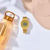 A gold Casio digital watch is placed next to a translucent amber water bottle and a beige baseball cap on a white background. Produktbild-Vorschau