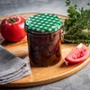 A jar of preserved tomatoes with a tomato and herbs on a wooden board. Produktbild-Vorschau