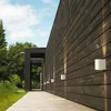 Exterior view of a modern wooden house with wall-mounted lights and a wooden walkway. Produktbild-Vorschau