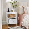 A bedside table with a lamp, vase, and basket next to a bed with pink and striped bedding. Náhľad obrázku produktu