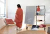 A person wrapped in a coral blanket stands next to a display rack with various home decor items, including blankets and pillows. Produktbild-Vorschau