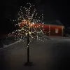 A decorative LED tree with warm white lights stands in the snow at night, with a red building in the background. Náhled obrázku produktu