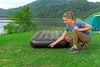 A woman is kneeling on grass next to an inflatable mattress near a lake and a tent. Náhled obrázku produktu