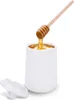 A white ceramic honey pot with a wooden honey dipper dripping honey into it. A white lid is placed next to the pot. Náhľad obrázku produktu