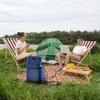 A couple camping by a river, with a tent, chairs, a cooler, and a small table with fruit. Náhled obrázku produktu