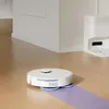 A white robot vacuum cleaner is shown mopping a section of a light wood floor. A white charging dock is visible in the background. Produktbild-Vorschau