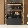 A modern display cabinet with glass doors and LED lighting, showcasing various items like bottles, dishes, and decorative objects. The cabinet is set against a textured stone wall and a wooden panel. Podgląd zdjęcia produktu