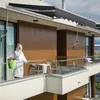 A woman in a white robe stands on a balcony, holding a cup and looking out. A dark gray awning is extended above her. Náhled obrázku produktu