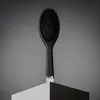 A black oval hairbrush with silver accents is displayed on a pedestal against a gray background. Produktbild-Vorschau