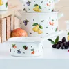A close-up shot of a small white pot with a fruit garden design, placed on a white surface. Several other pots are visible in the background, along with some fruit and grapes. Produktbild-Vorschau