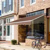 A brown retractable awning is installed over a storefront window, with a bicycle parked nearby on the sidewalk. Produktbild-Vorschau