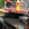 A person is grilling meat on a large, round grill with flames erupting from the meat. Produktbild-Vorschau