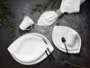 A flat lay of white porcelain dinnerware set on a dark textured surface, with green sprigs and a white napkin. Produktbild-Vorschau