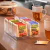 A clear acrylic tea box filled with various flavors of Pickwick tea bags sits on a wooden table next to a glass mug of tea and a jar of cookies. Produktbild-Vorschau
