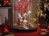 A festive Christmas display featuring a glass cloche with ornaments, berries, and pinecones, set on a red tablecloth with a Christmas tree in the background. Produktbild-Vorschau