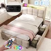 A bedroom scene featuring a beige upholstered bed with storage drawers, a remote control for LED lights, and a close-up inset of electrical outlets and USB ports. Produktbild-Vorschau