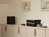 A black Denon CD player and amplifier are stacked on a white cabinet, with a black subwoofer to the left and stacks of CDs on the right. Two framed abstract art pieces hang on the wall above. Produktbild-Vorschau
